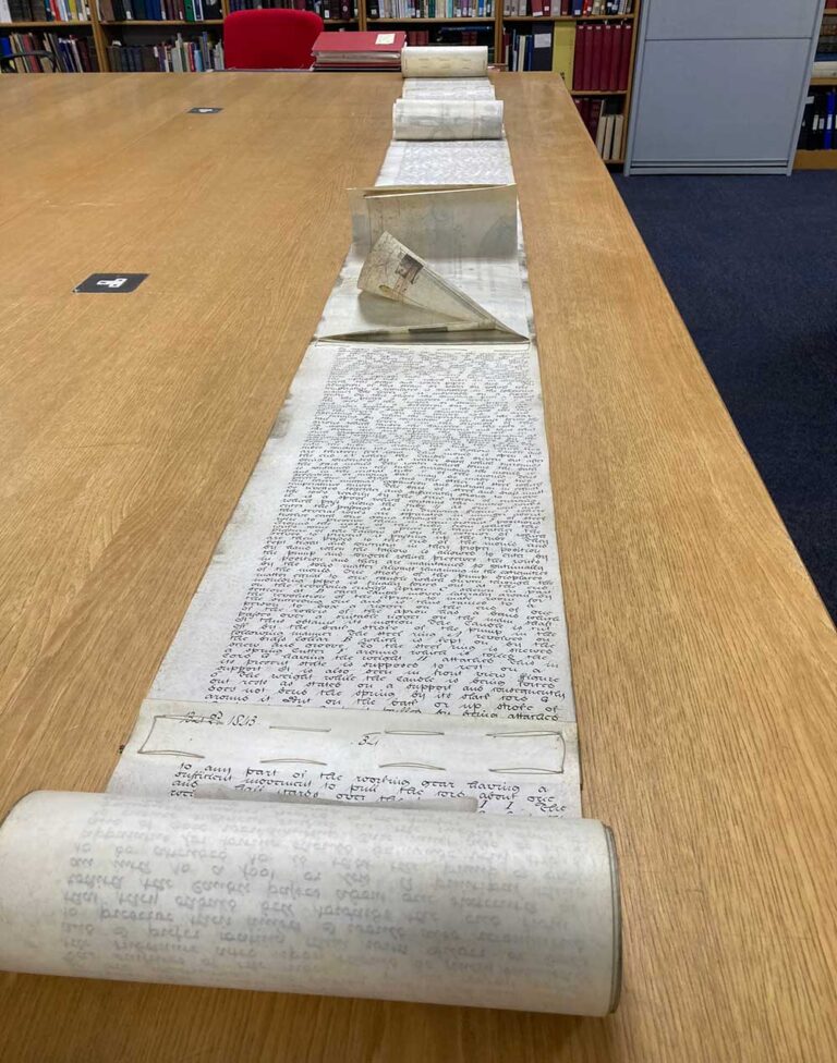 A very long roll of hand written document, partially unrolled. It is the full length of a table. You can see where pieces of paper have been sewn together to make the roll. 
