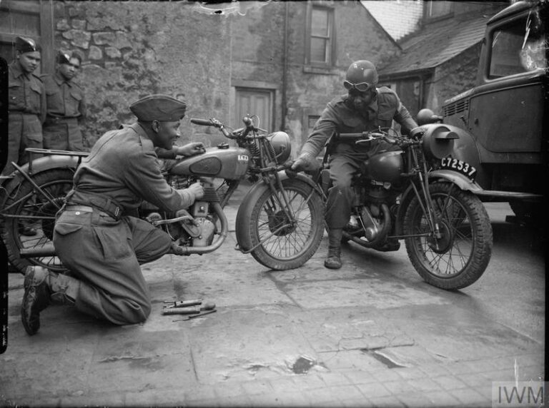 A black-and-white photograph of two West Indian troops are talking, one sitting on a motorbike the other seemingly fixing a motorbike with mechanic tools next to him. 