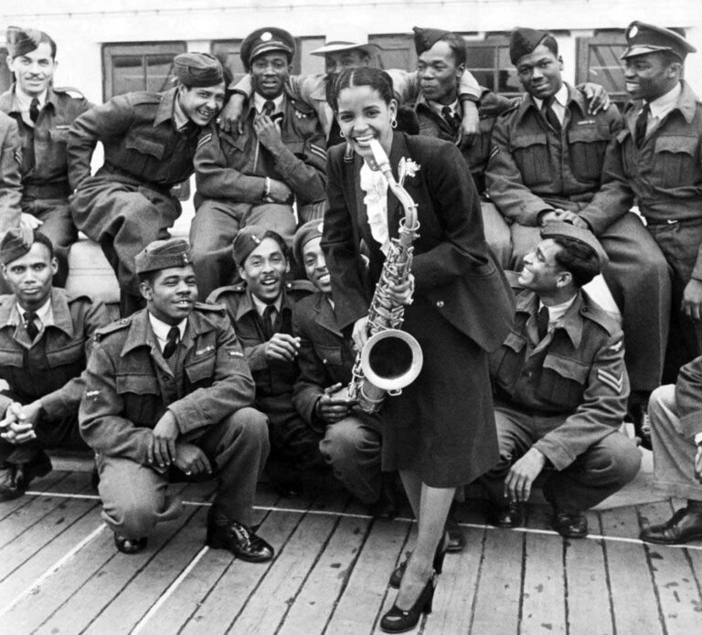 A young woman holding a saxophone smiles in front of two rows of uniformed young men.