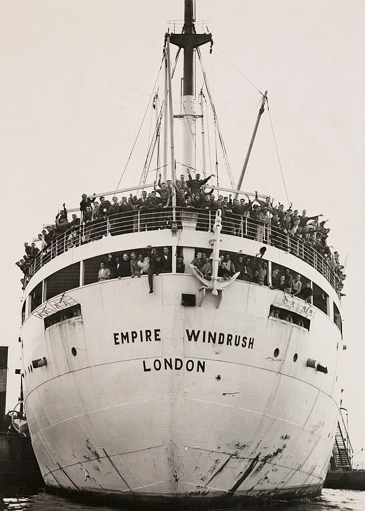 A boat is docked in a port. The words 'Empire Windrush London' is written on the hull. Passengers on the deck and are waving. 