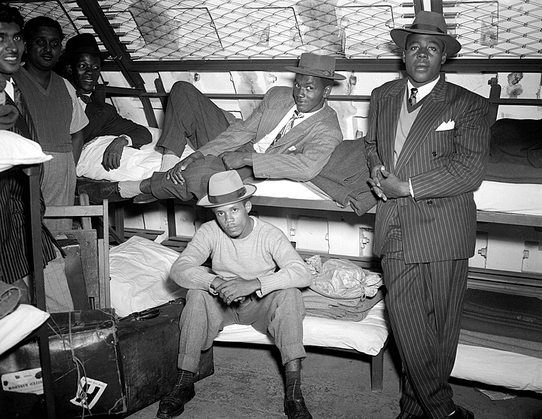 6 Windrush passengers sit, lie and stand next to bunkbeds in the Clapham South shelter while be photographed. They are wearing smart clothing including suits and trilbies and looking towards the camera. Luggage sits besides them.