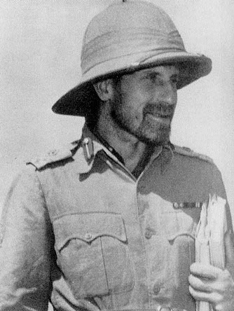 A bearded person wearing a military hat and shirt and holding some papers.