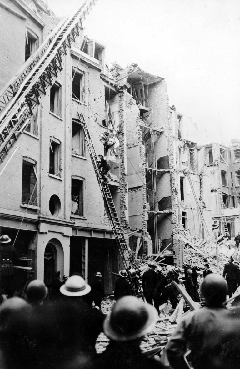 Workers in helmets and members of the public watch a figure climbing a ladder to the fourth storey of a damaged building.