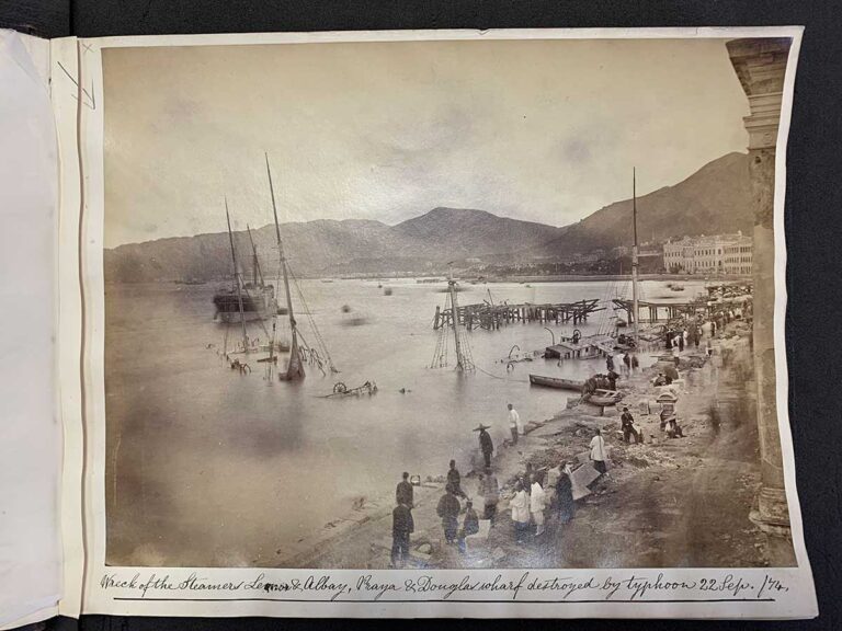 A sepia photo showing around a dozen people stood on land beside a sunken ship, with its deck just underwater.