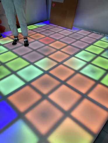 A person is stood on a interactive board  designed in squares. Each square is lit up with a bright colour, from blue, green, and red.  