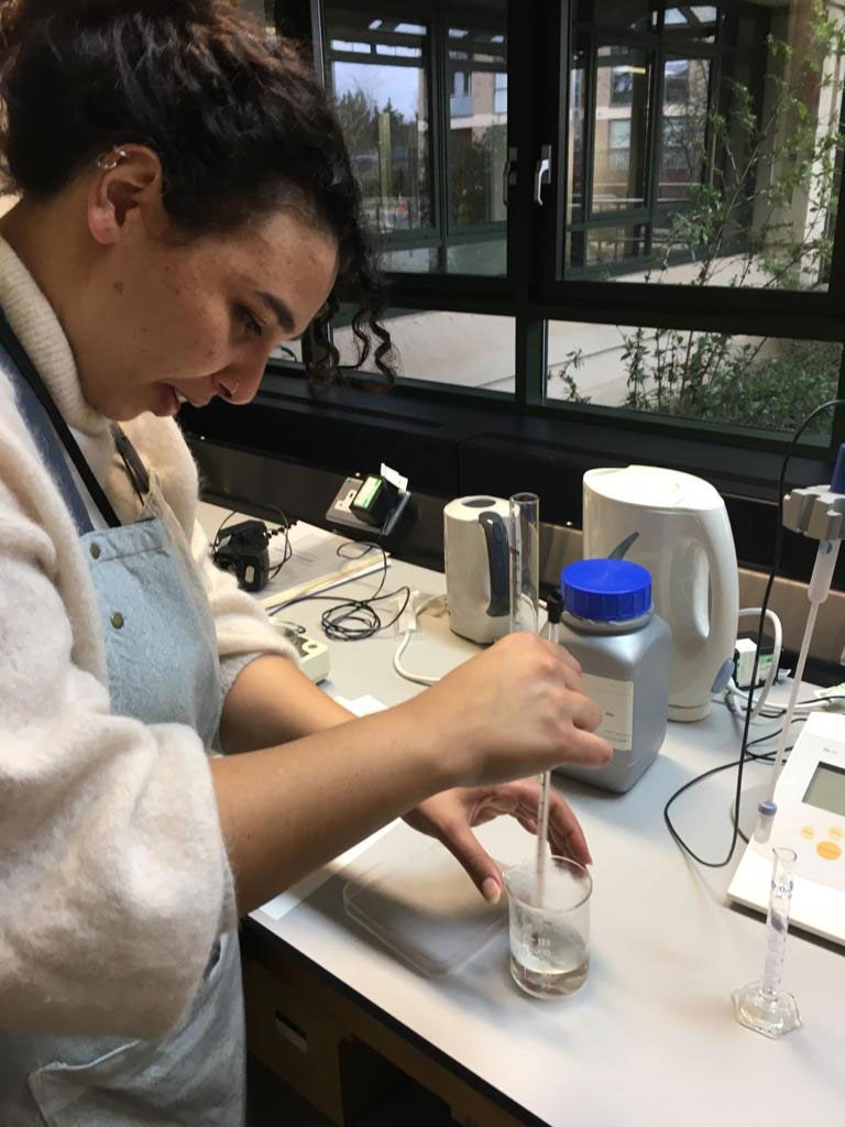 A conservator stands at a bench holding a thermometer in a small glass jar holding a clear liquid. 