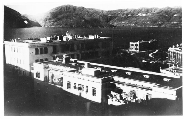 The outside of a wing of Stanley Civilian Internment Camp. It sits by the sea and there are mountains surrounding it. 