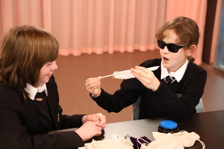 A student holds a quill with a surprised expression. They sit with another student next to a table with different clues from the hint bags used during the workshop.