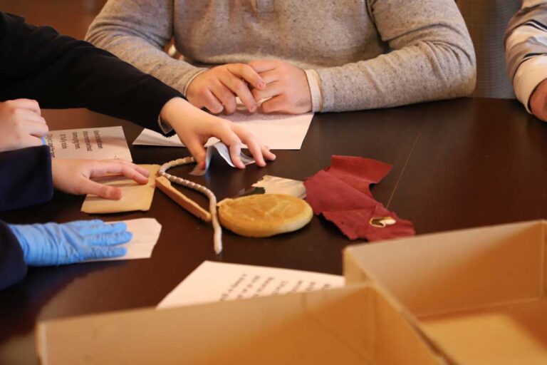 Students sit at a table with archival samples including snake weights and a wax seal, as well as worksheets in large print and Braille.