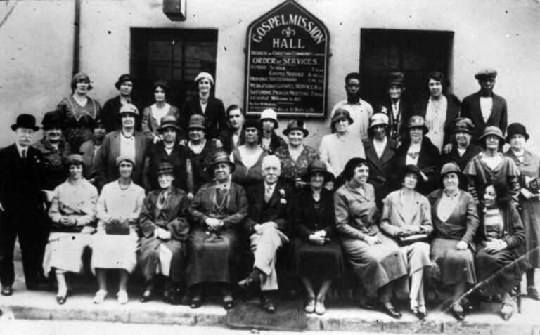 Photo of three rows of people, including two West African men, around a sign saying 'Gospel Mission Hall'.