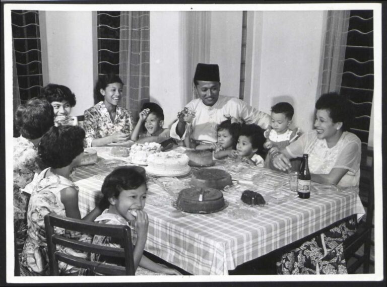 Photograph of A Malay family of adults and children sitting around a dining table celebrating the end of fasting with cakes and treats.