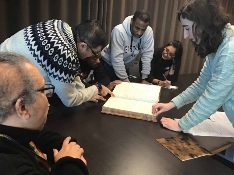 A group of visitors at The National Archives stand and sit at a table looking at a book.