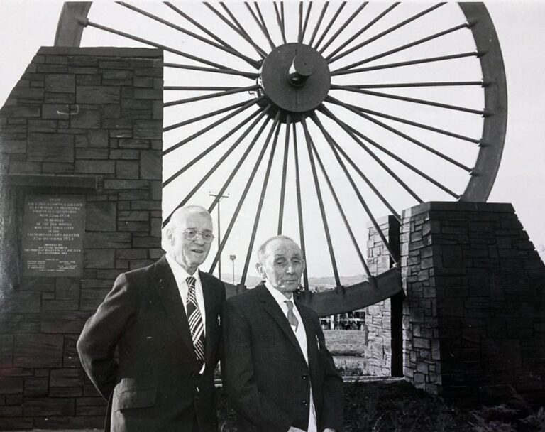 Two elderly men wearing suits standing in front of a large wheel from a coal pit, now used as a memorial. 