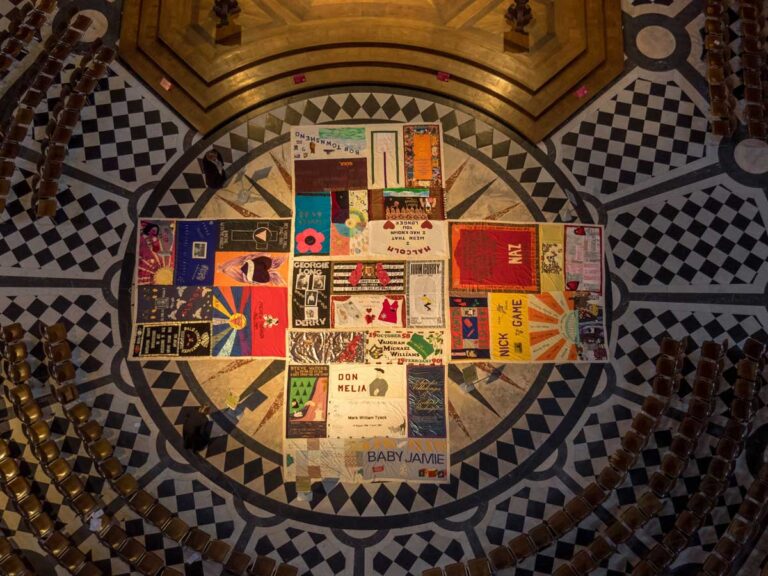 Dozens of quilts stitched together and lying in a cross shape on the floor in the middle of a ring of chairs.