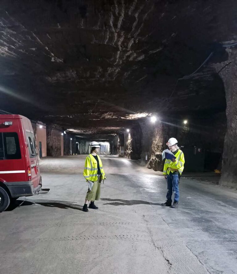 Two staff members of The National Archives stand in the DeepStore salt mine wearing hard hats and Hi-Vis jackets. 