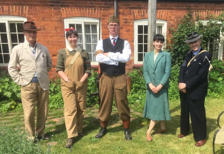 Five people stand in a garden looking to the camera, some wearing smart suits, the others in practical clothes like dungarees.