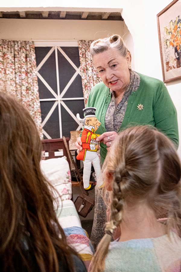 A person with her silver hair curled holds a stuffed doll marked 'Sonny Jim' and talks to two children.