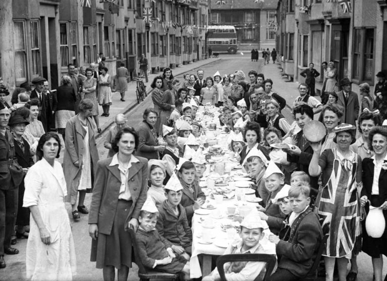 Black and white photograph of dozens of children wearing party hats, sat at one long table in the middle of a street covered in Union Flag bunting.