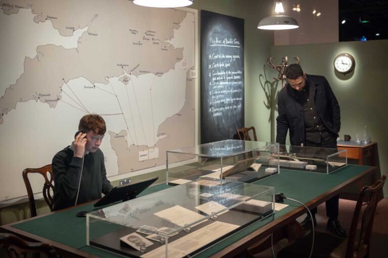 One exhibition visitor sits at a table with a speaker to her ear, while another looks down at a glass display case filled with historical documents, in front of a large map of the English Channel.