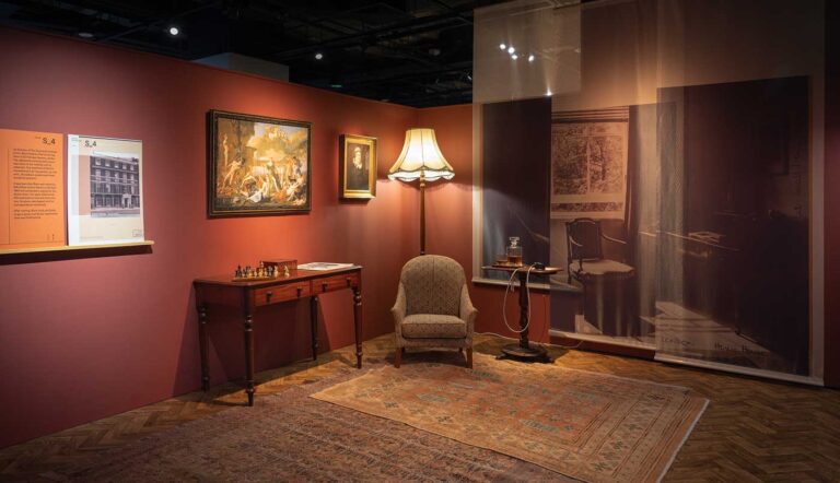 A cushioned armchair sits next to an ornate wooden table at the centre of a faded rug, beside a hanging showing Anthony Blunt's real flat.