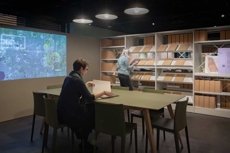 A woman sits at large table and lifts a sheet of paper from an opened file, in front of shelves of others and a map projected on to the wall.